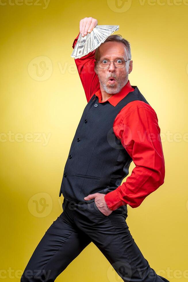 Old smiling grey-haired man in eyeglasses holding fan of dollars over head in dancing pose. Human emotions and facial expressions photo