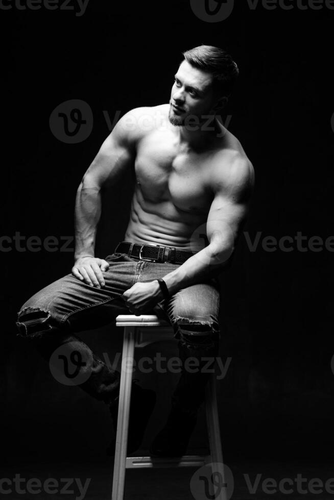 Muscular and fit young bodybuilder fitness male model posing on chair. Black and white photo. Full size portrait. photo