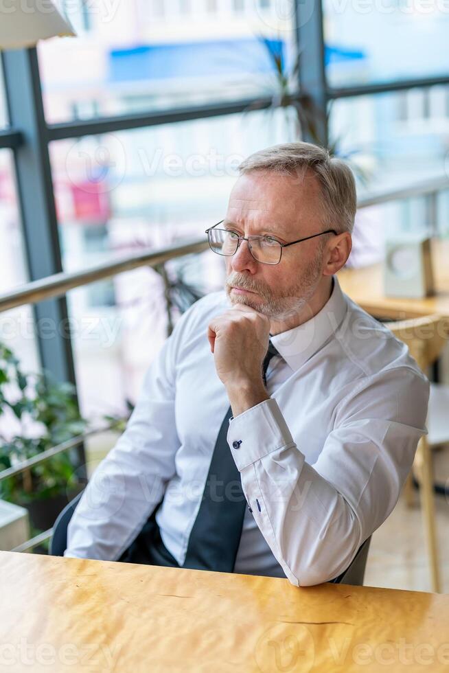 Mature businessman looking in front. Thinking over ideas. Sitting near the window in cafe. Panoramic city view background. Business photo. photo