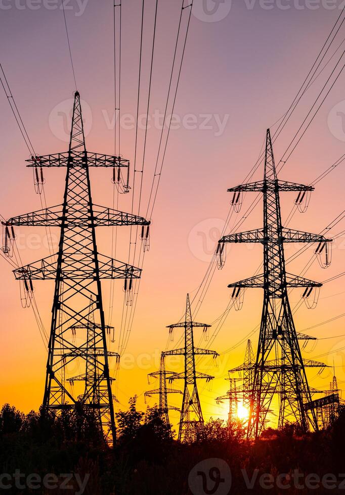 High voltage electric towers at golden sunset. Silhouettes of a number of electricity pylons and trees. Power towers at the background of bright setting sun. photo