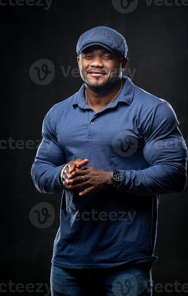 Confident smiling man holds hands crossed. Photo of african man in casual outfit on black background.