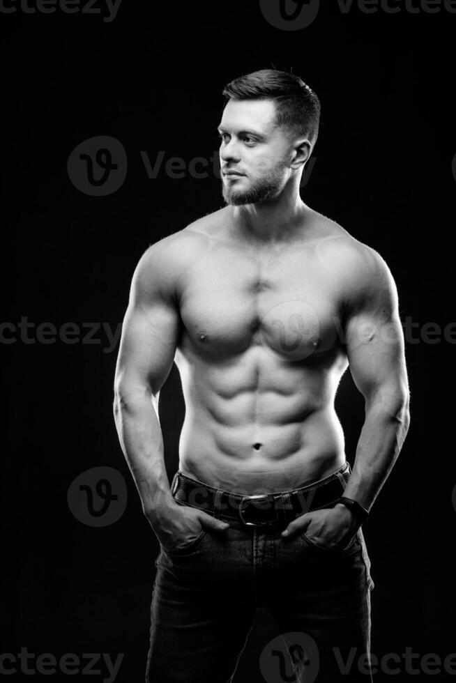 A portrait of a hot handsome strong guy man without a shirt against dark background. hands in pockets. Studio photo. Male beauty concept. Black and white. Closeup. photo