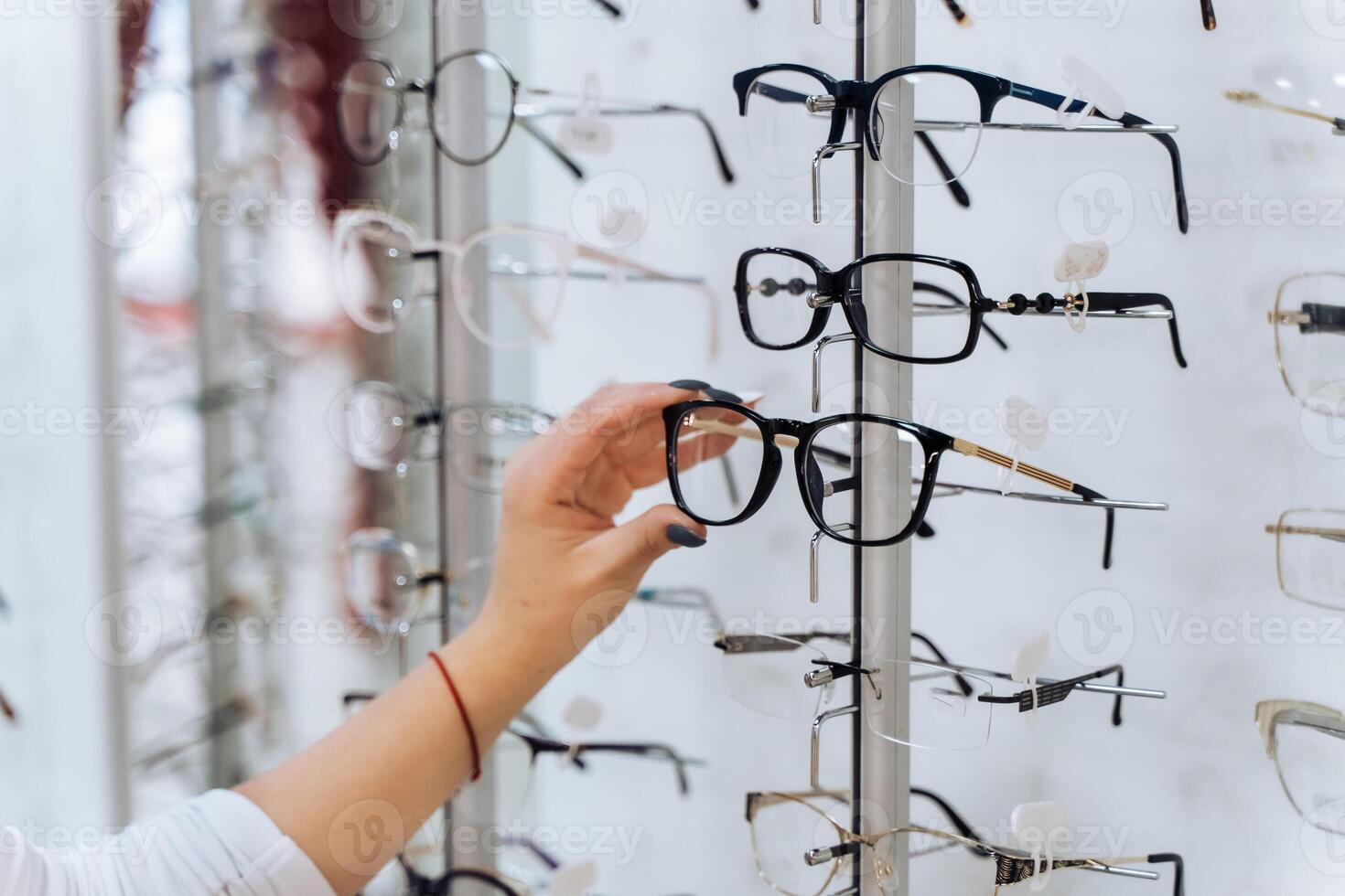 Choosing glasses. Client in optics. Selective focus on hand. Woman touching corrective glasses on stand with spectacles. Wide assortment in shop. photo