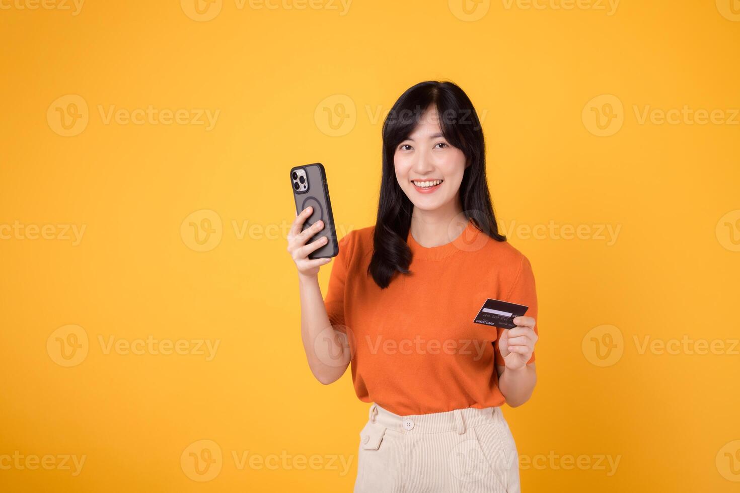 Thrilled young Asian woman 30s in orange shirt, using smartphone and holding credit card on yellow background. Delightful online shopping. photo