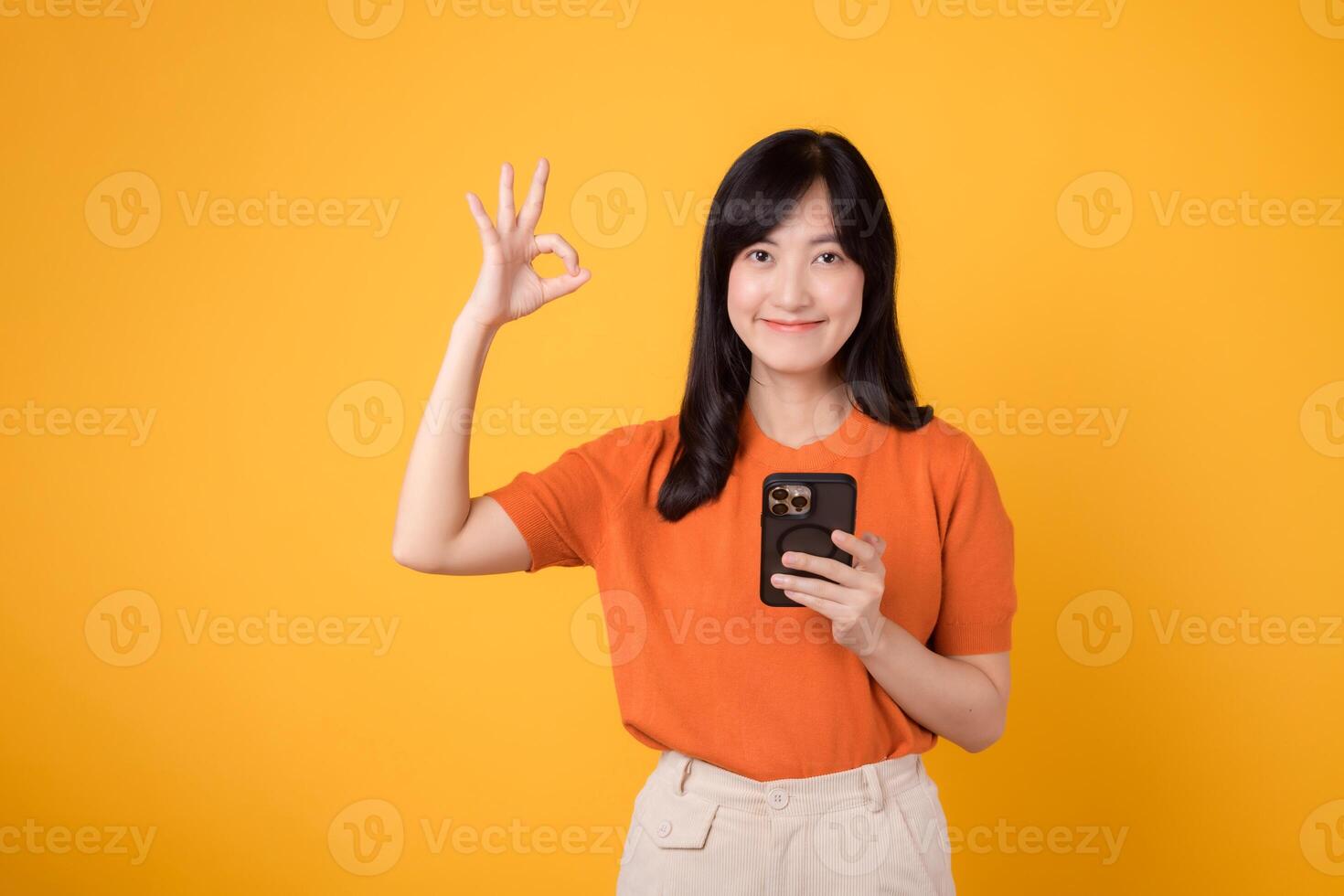 Cheerful Asian woman in her 30s, wearing orange shirt, using smartphone with okay hand sign on vibrant yellow background. New mobile app concept. photo