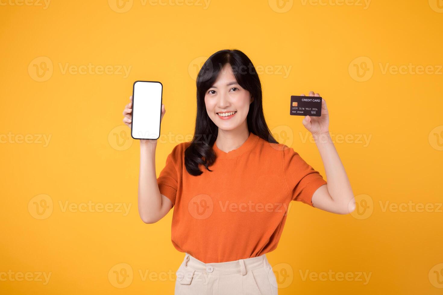 Excited Asian woman in her 30s, presenting smartphone and credit card on yellow background. Convenient online payment shopping. photo