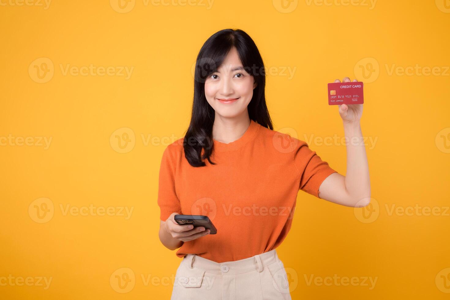 Vibrant Asian woman in her 30s, using smartphone and showing credit card on yellow background. Effortless online shopping. photo