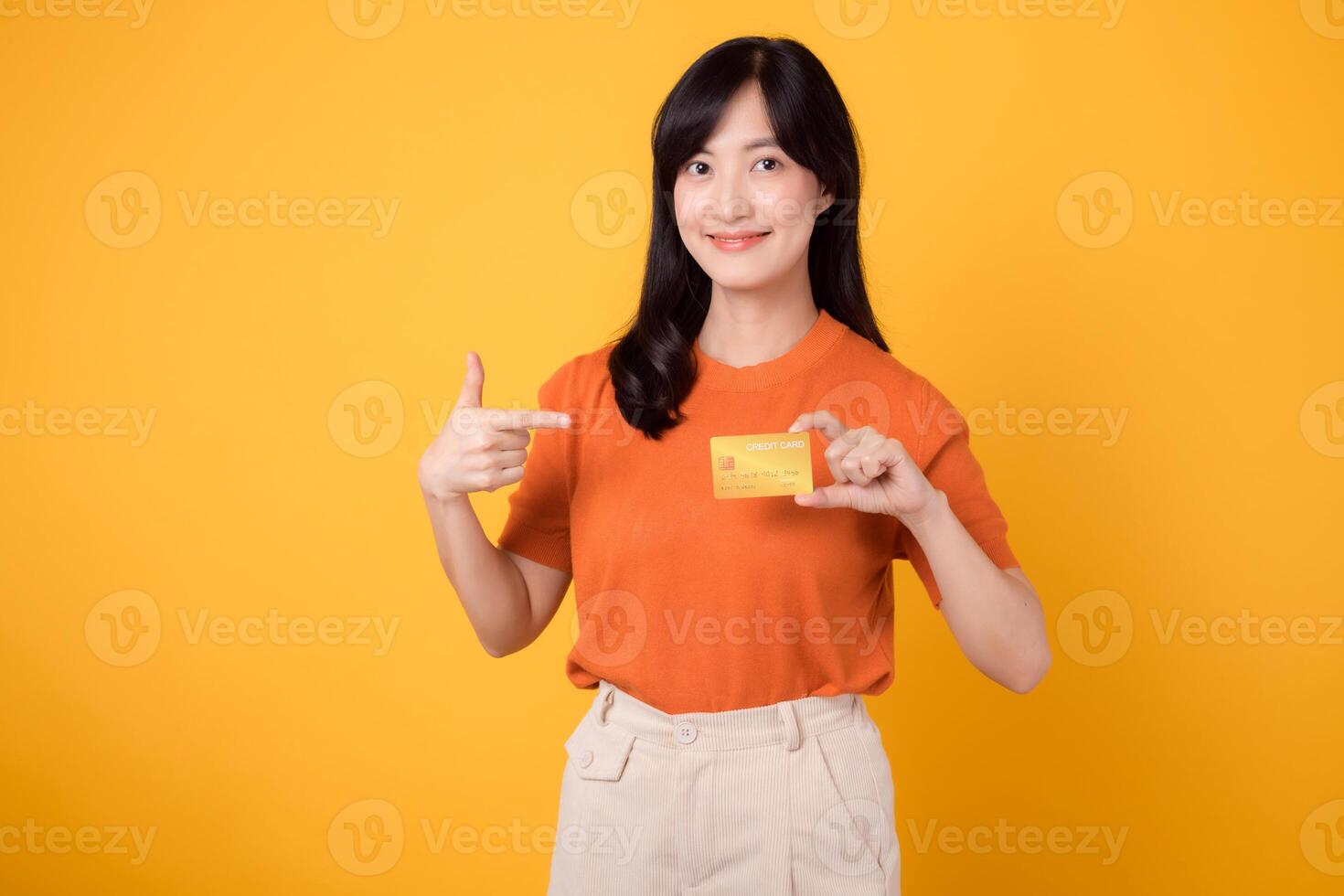 Young Asian woman in her 30s wearing orange shirt, pointing finger to credit card on vibrant yellow background. fast online shopping payment shopping. photo