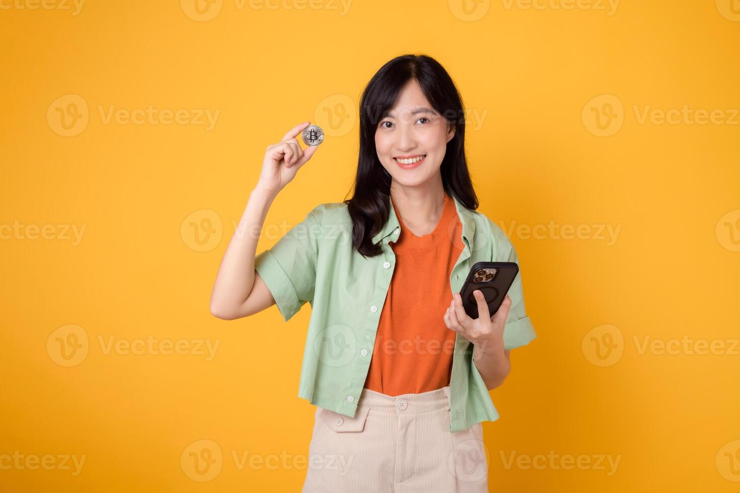 joven asiático mujer en su 30, esmeradamente vestido en naranja camisa y verde saltador, utilizando cripto moneda moneda mientras participación teléfono inteligente en amarillo antecedentes. futuro Finanzas concepto. foto