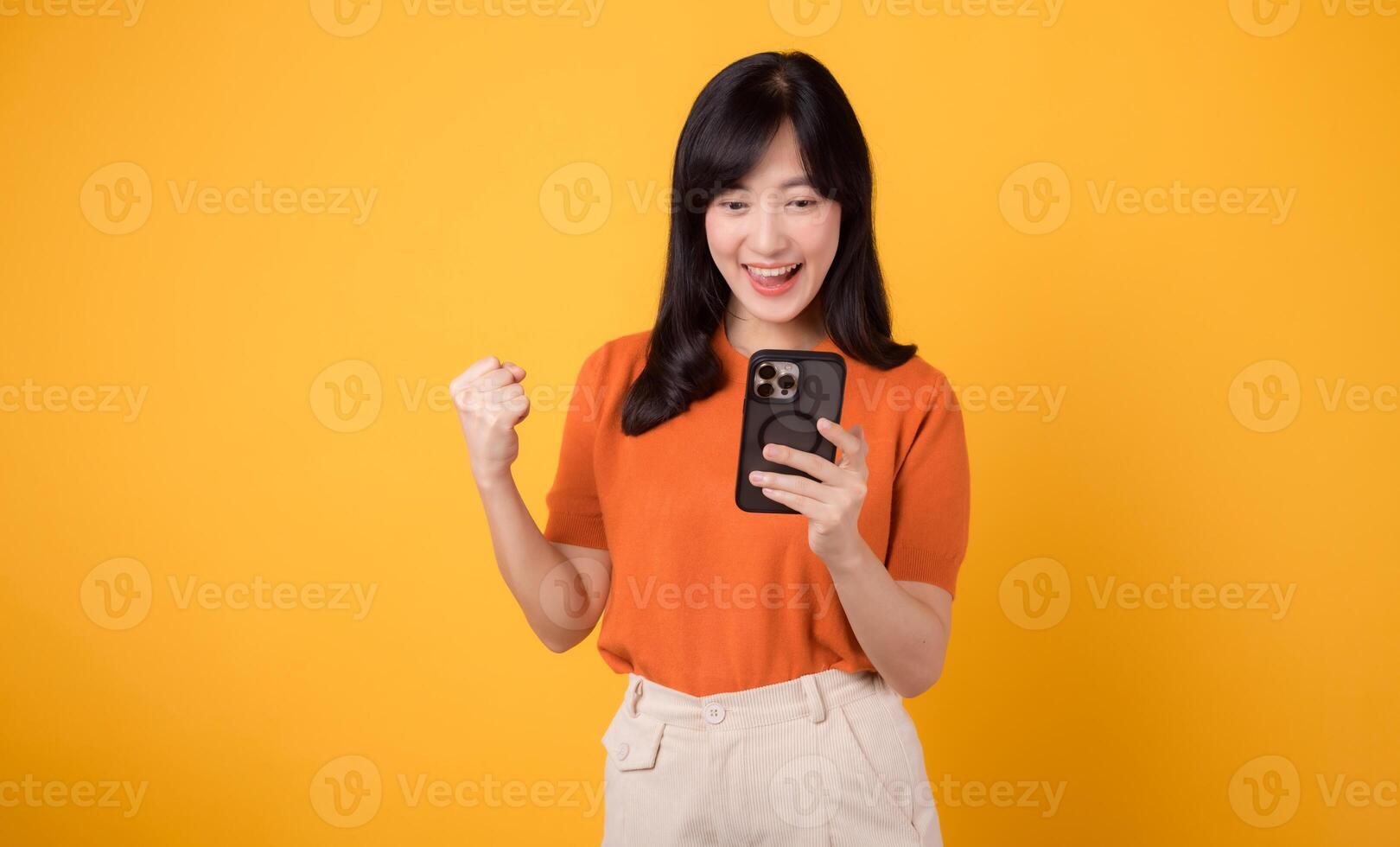 Joyful Asian woman in 30s, wearing orange shirt, using smartphone with fist up hand sign on vibrant yellow background. Celebrate new app concept. photo