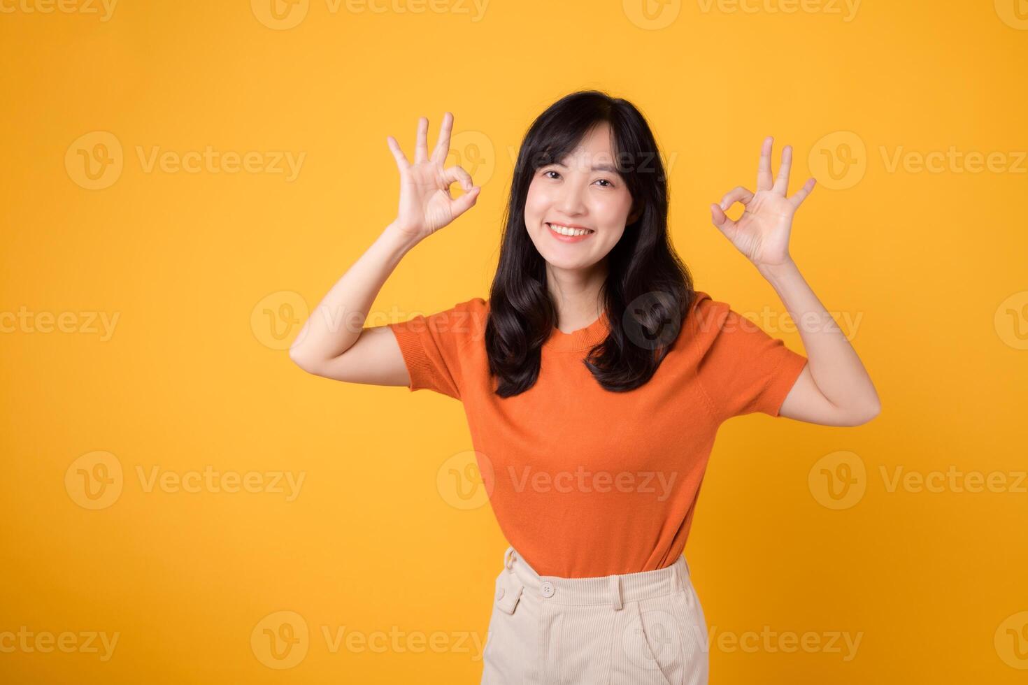 A young Asian woman in her 30s, wearing an orange shirt, showcases the okay sign gesture on a sunny yellow background. Hands gesture concept. photo