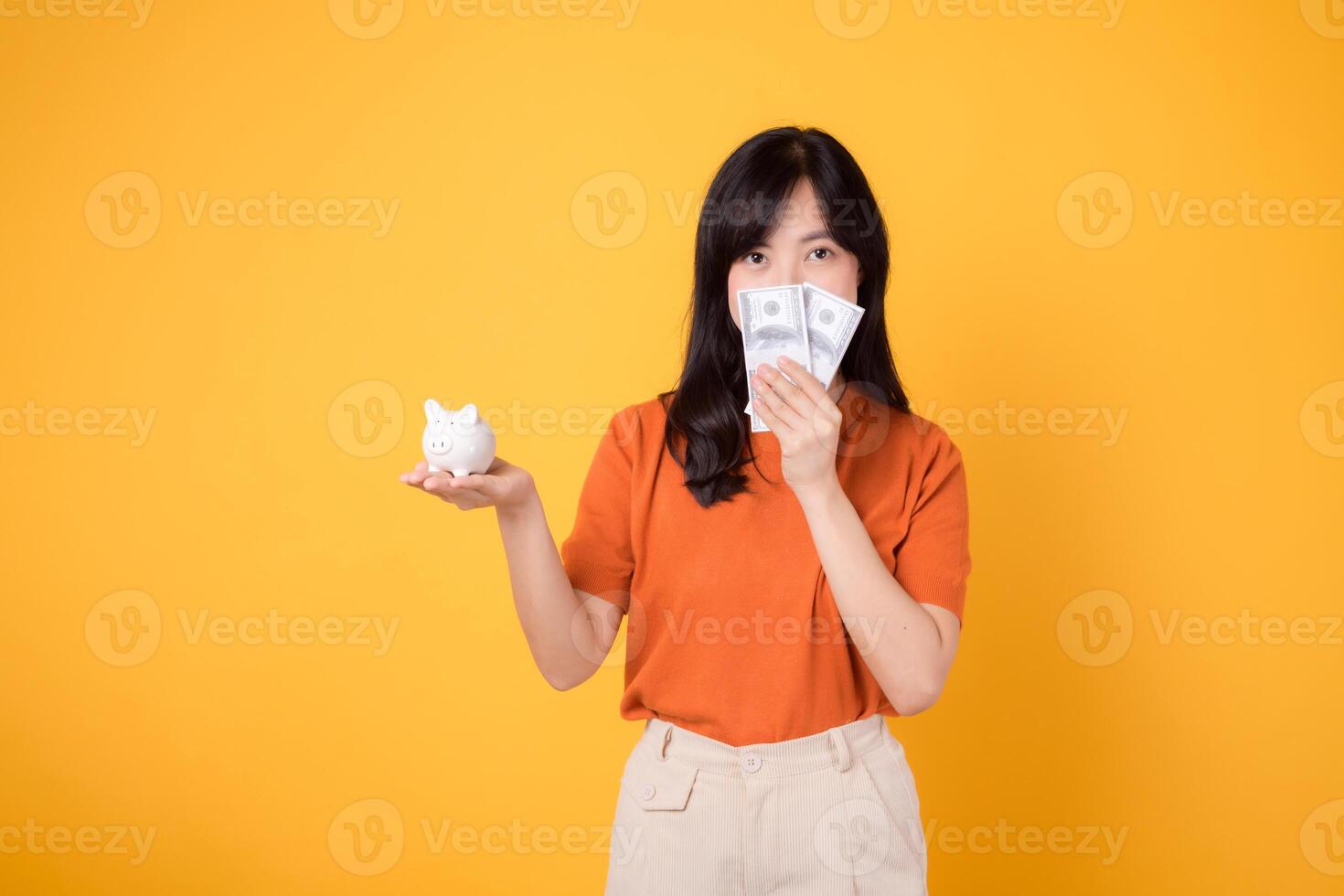 Joyful Asian woman in her 30s kisses piggy bank, holds cash money dollars on vibrant yellow background. Wealth saving concept. photo