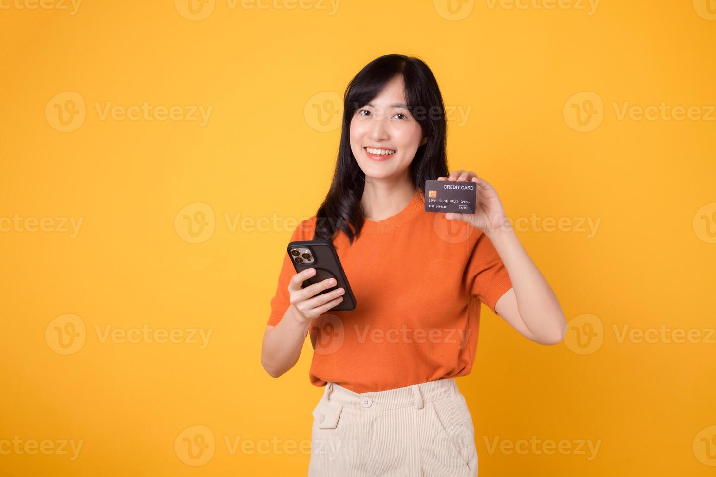 Vibrant young Asian woman in her 30s, using smartphone and holding credit card on yellow background. Effortless online shopping. photo