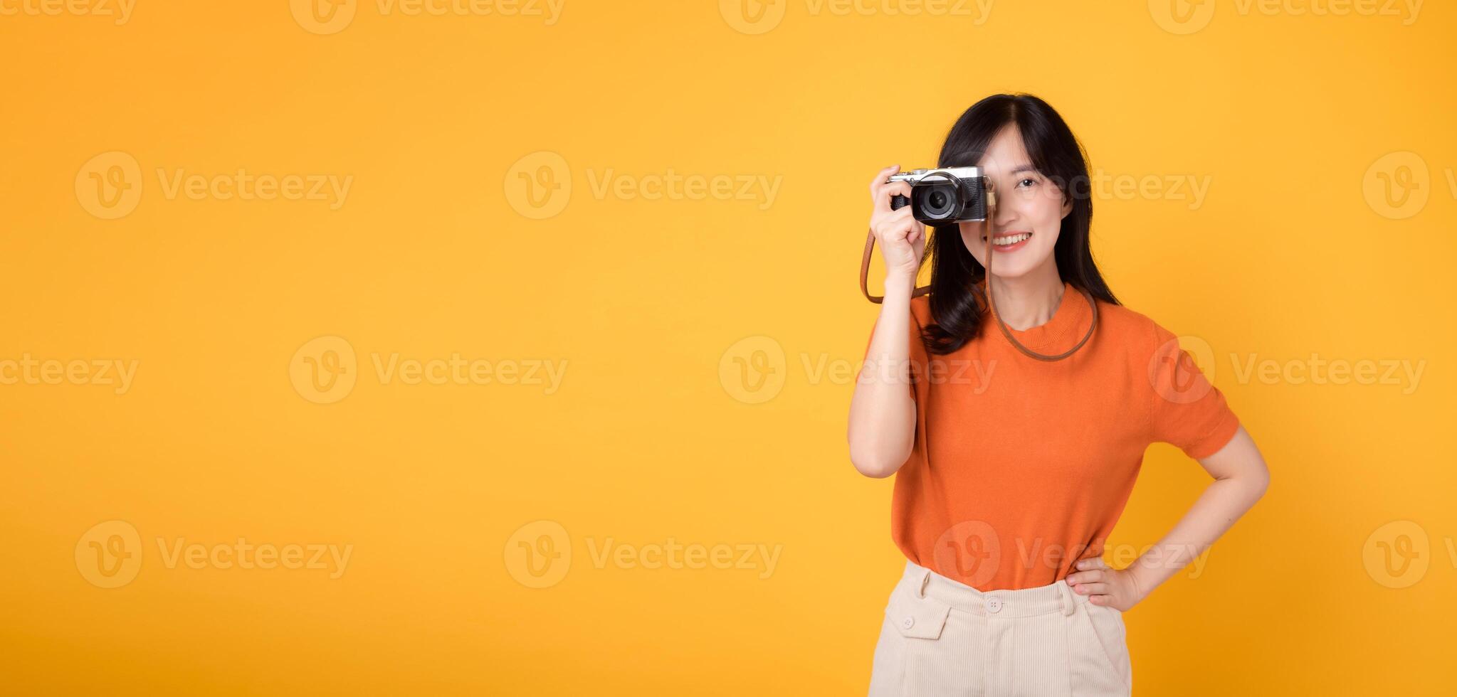 Excited woman traveler capturing holiday memories with a camera isolated on yellow background. Adventure concept. photo