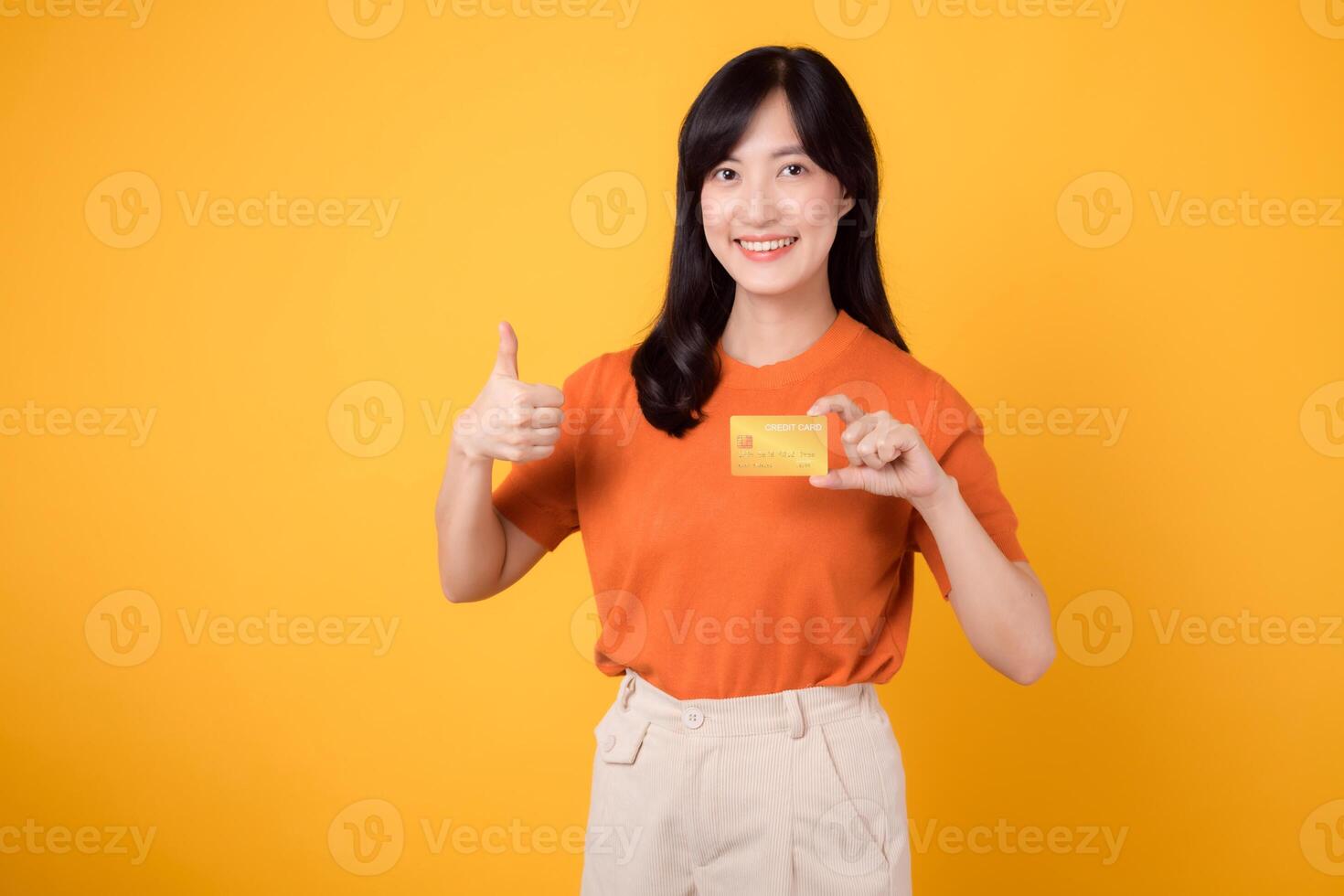 Confident young asian woman 30s holding a credit card, standing isolated on vibrant yellow background. Secure online payment concept photo