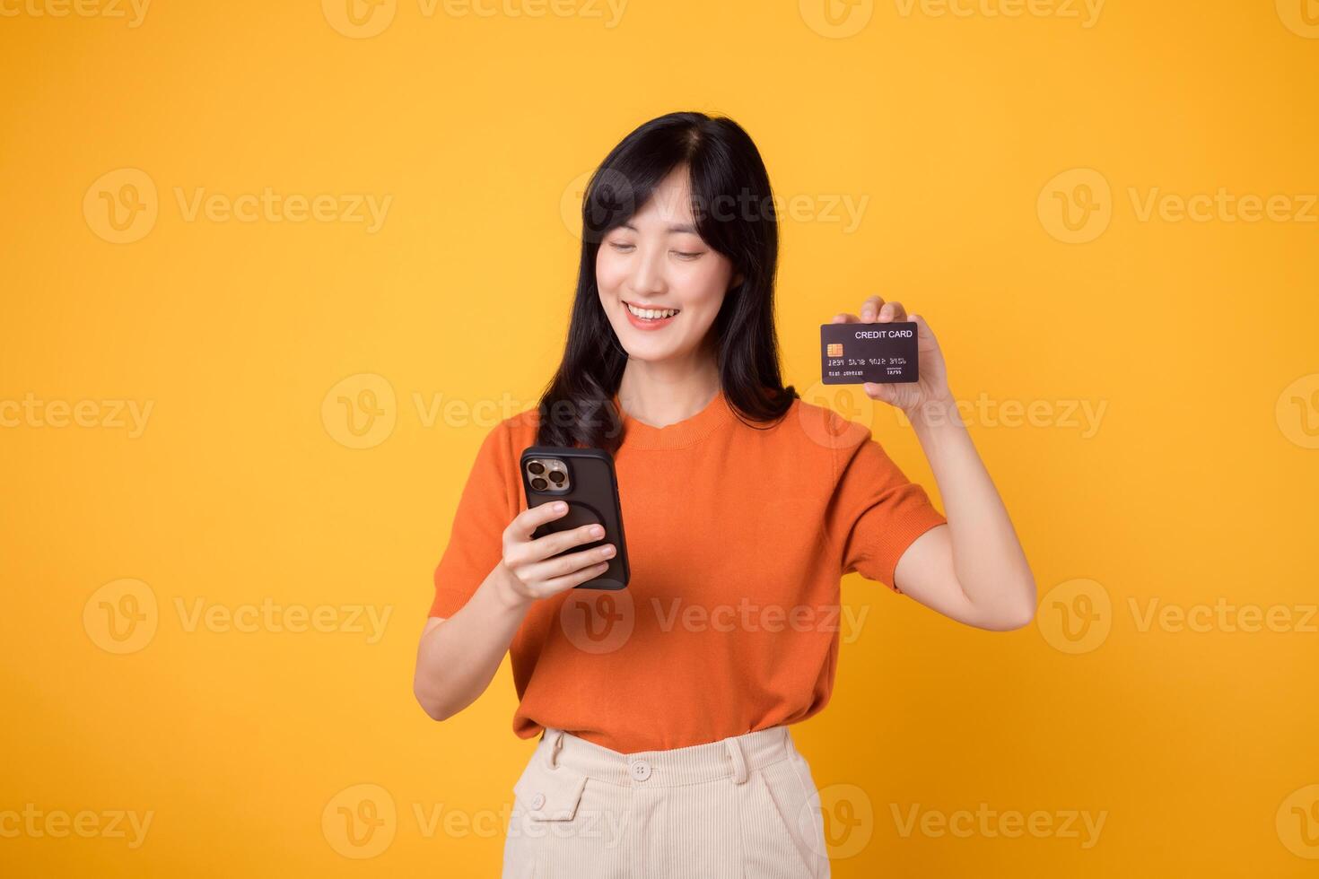 Enthusiastic Asian woman in her 30s, using smartphone and showing credit card on yellow background. Swift online shopping. photo