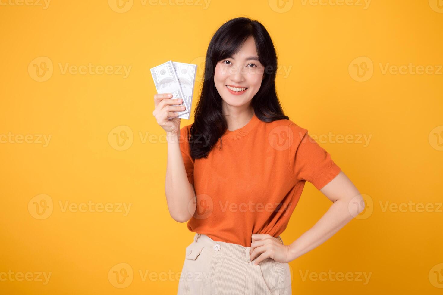 Dynamic young Asian woman in her 30s, holding cash money dollars, standing on vibrant yellow backdrop. photo