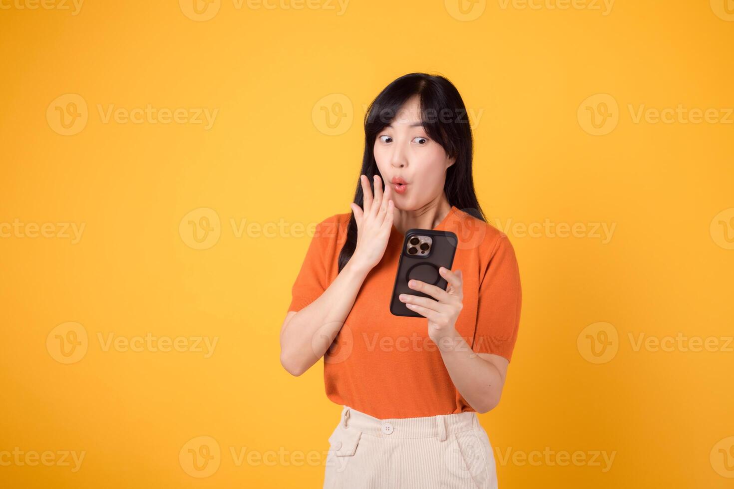 emocionado asiático mujer en su 30, vistiendo naranja camisa, utilizando teléfono inteligente con puño arriba mano firmar en vibrante amarillo antecedentes. emocionante nuevo aplicación experiencia. foto