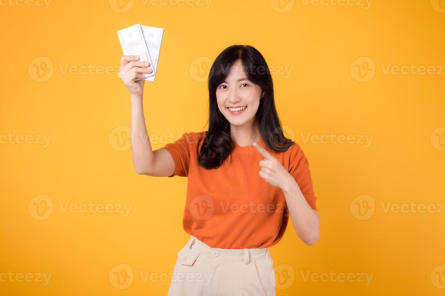 Dynamic Asian woman in her 30s points to cash money dollars on vibrant yellow backdrop. Wealth and prosperity concept. photo