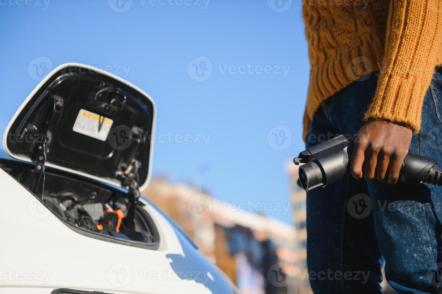 Close up of african american man connecting charging cable to electric car. Young male standing near his modern auto with leather suitcase in hand. photo