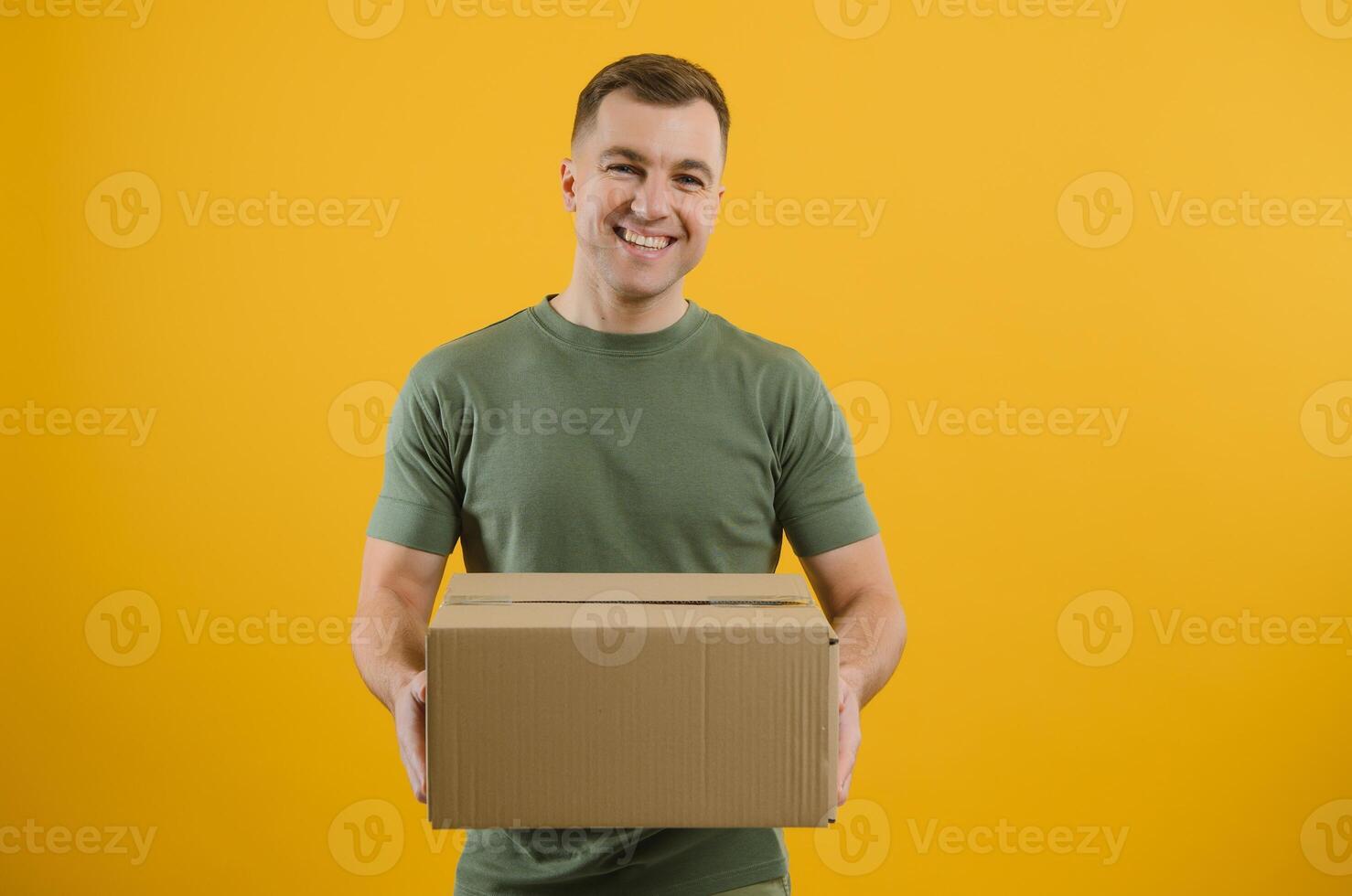 Delivery man in green uniform isolated on yellow background, studio portrait. Male employee working as courier dealer hold empty cardboard box. Service concept. Mock up copy space photo