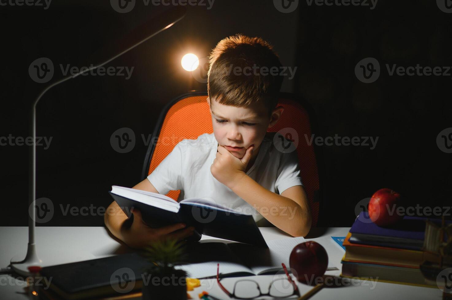 concentrado colegial leyendo libro a mesa con libros, planta, lámpara, color lápices, manzana, y libro de texto foto