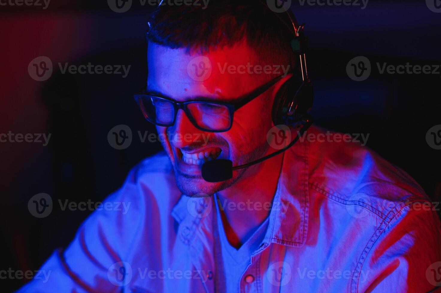 Professional Gamer Plays Video Game on His Computer. He's Participating in Online Cyber Games Tournament or in Internet Cafe. He Wears Glasses and Talks into Microphone. photo