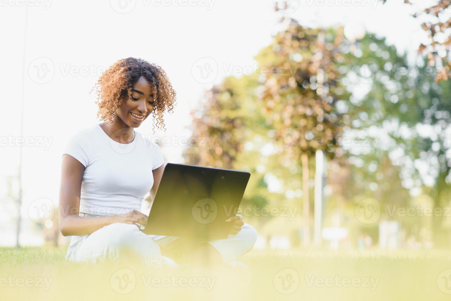 Thoughtful cute mixed female international student with curly hair is sitting on fresh grass with modern laptop in public park, leaning on apple tree and wistfully looking aside during her break photo