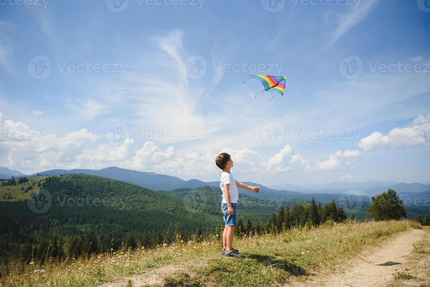 Little school age boy running down the slope with kite in the sun. Sunny summer or spring day at sunset. Active outdoor games and leisure. Mountain landscape with hills and coniferous trees. photo