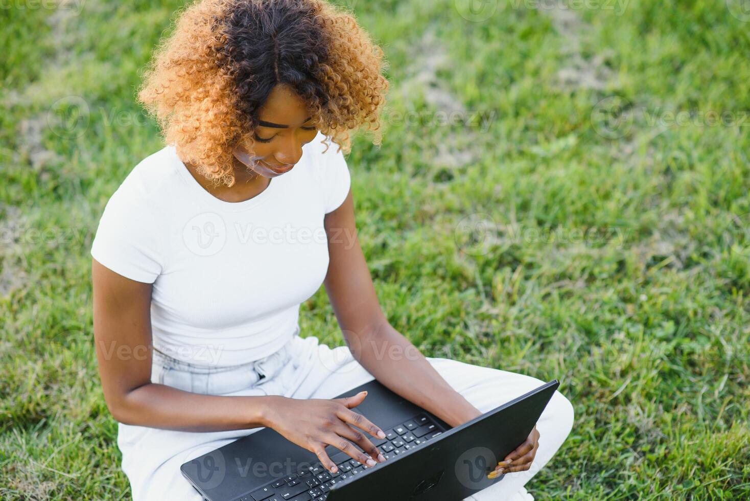 Human and technology concept. Charming African woman with short pixie hairstyle enjoying sunny weather, sitting on the lawn in front of laptop computer in the public garden, waiting for her friend photo