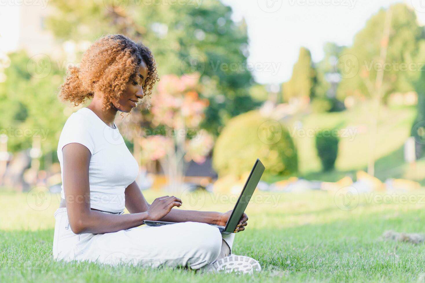 Thoughtful cute mixed female international student with curly hair is sitting on fresh grass with modern laptop in public park, leaning on apple tree and wistfully looking aside during her break photo