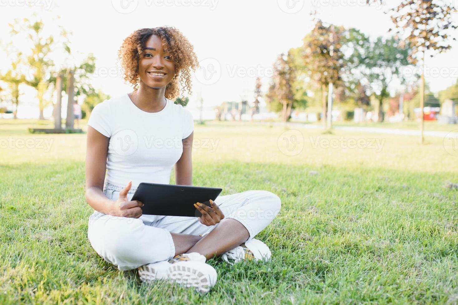 Thoughtful cute mixed female international student with curly hair is sitting on fresh grass with modern laptop in public park, leaning on apple tree and wistfully looking aside during her break photo