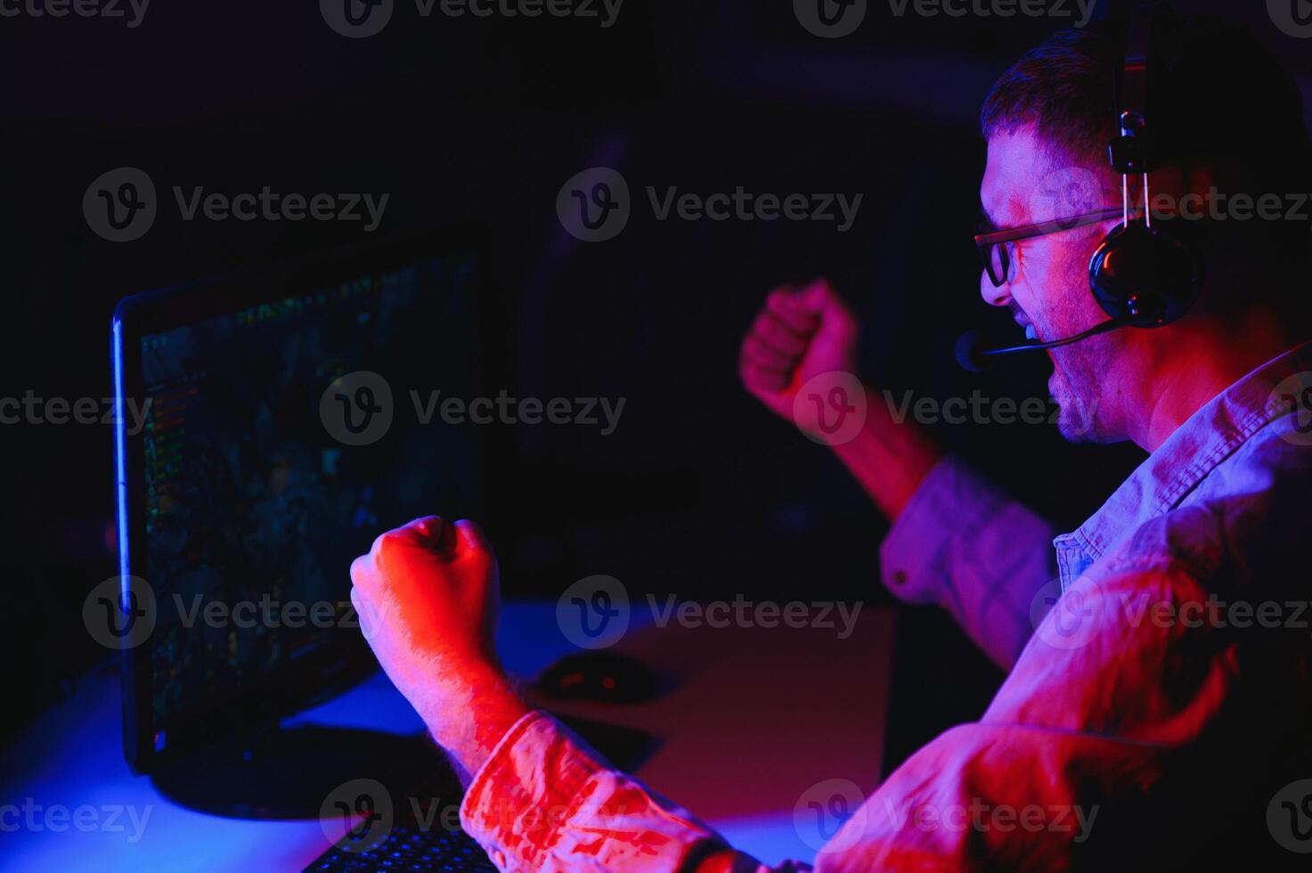 Professional Gamer Plays Video Game on His Computer. He's Participating in Online Cyber Games Tournament or in Internet Cafe. He Wears Glasses and Talks into Microphone. photo