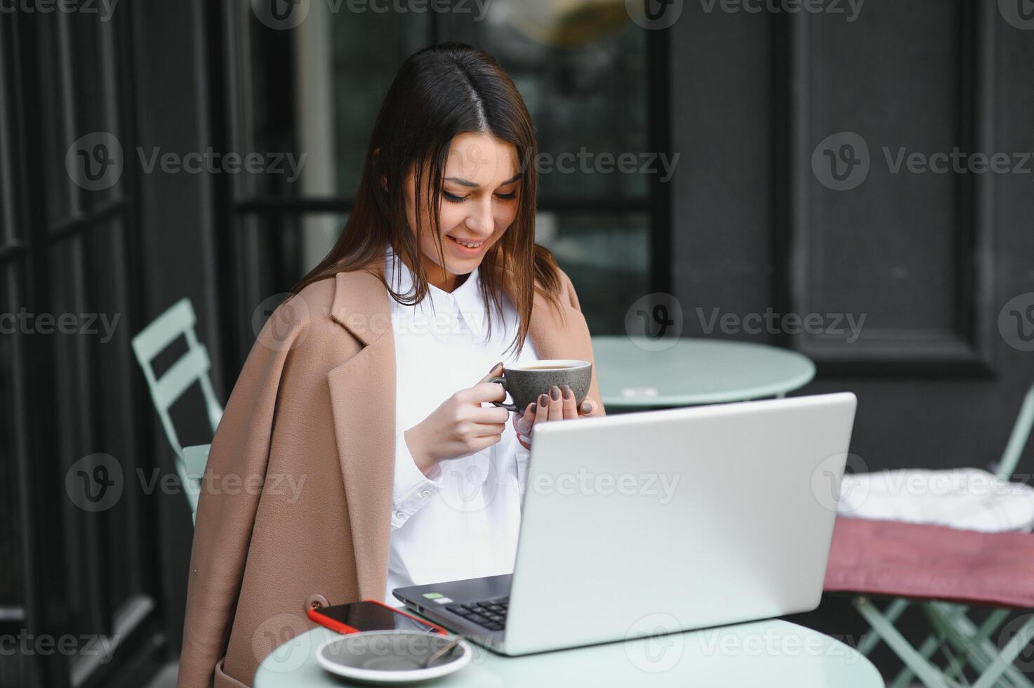 hermosa y contento mujer se sienta con un ordenador portátil en un café en el calle y bebidas café. Lanza libre trabajar. al aire libre reuniones foto