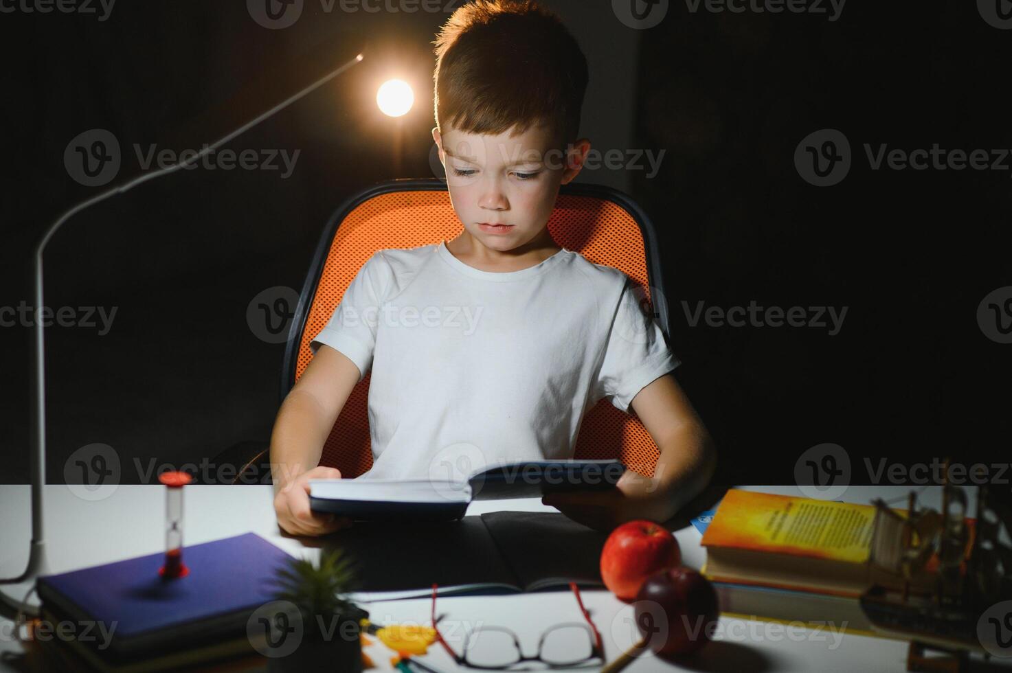 concentrated schoolboy reading book at table with books, plant, lamp, colour pencils, apple, and textbook photo