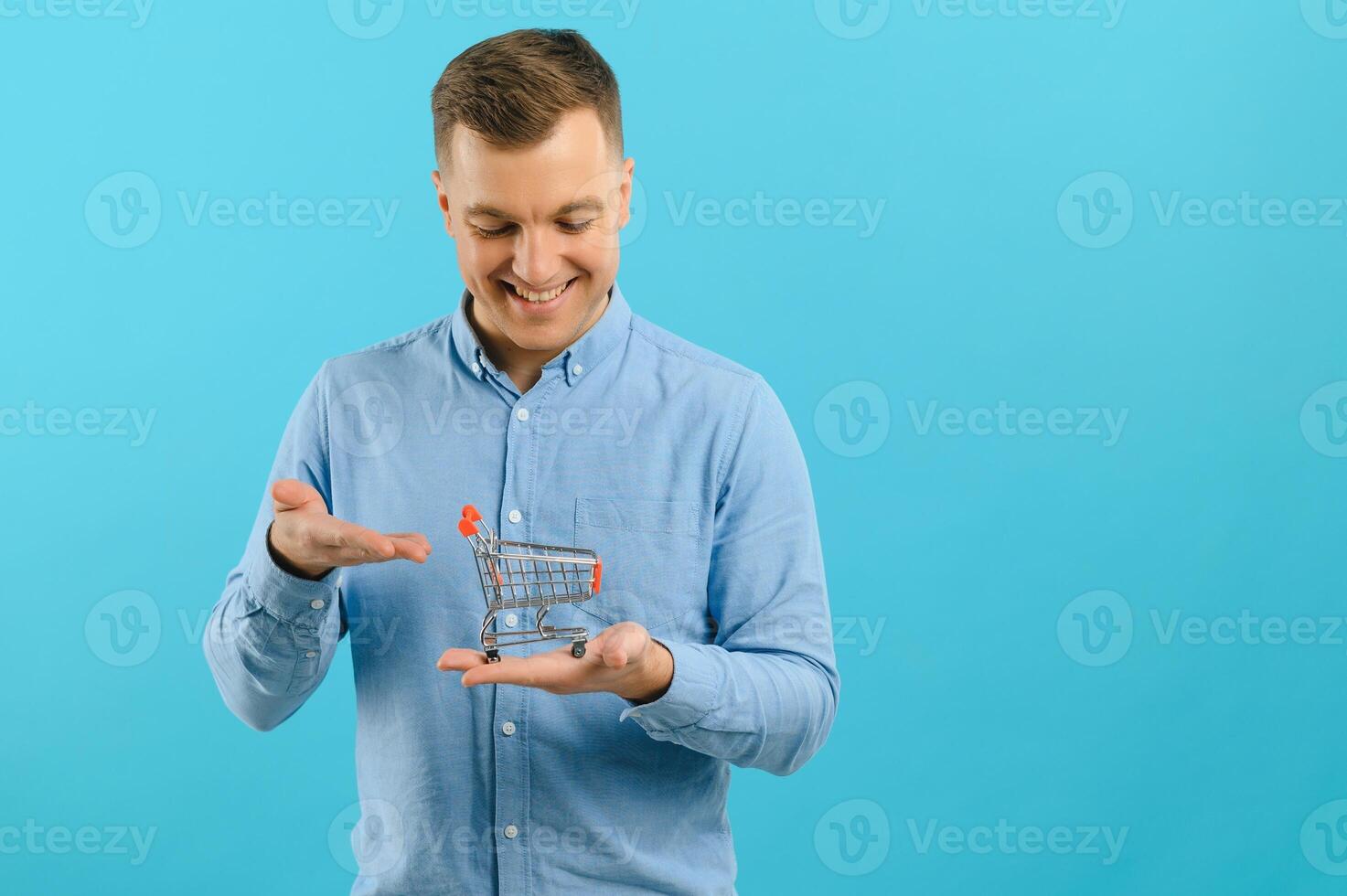 A young man holding a mini shopping cart and standing in front of a blue background in the studio. Sales concept. Place for text. photo