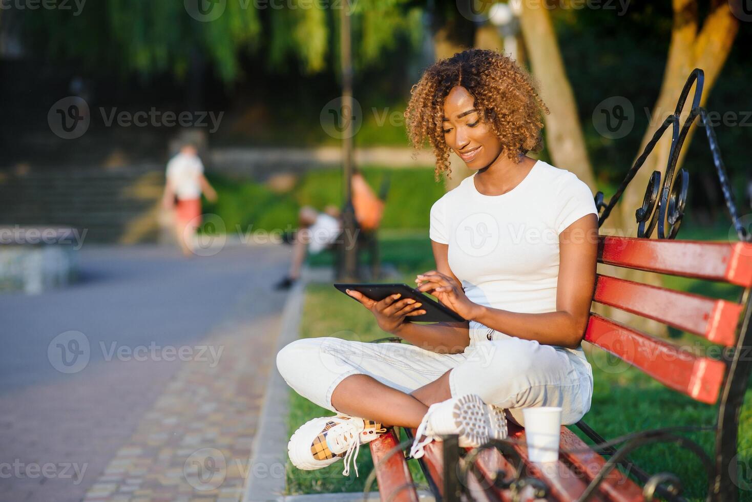 Young beautiful Brazilian female with black curly Afro hair with tablet, while sitting outdoors on the wooden bench in a park. photo