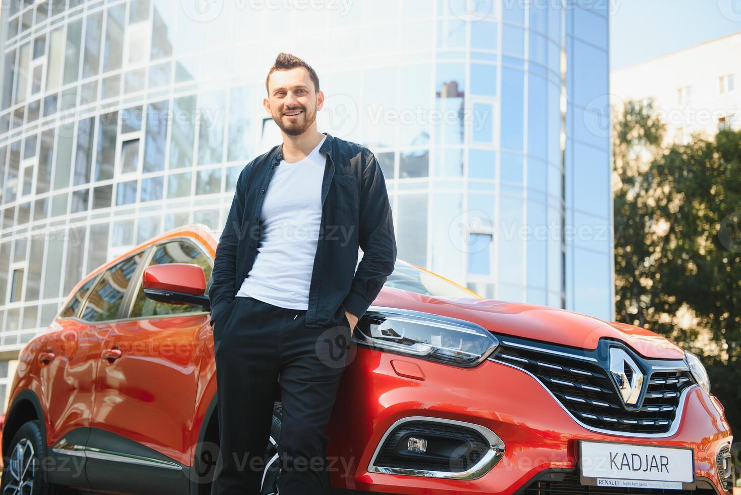 An attractive man with a beard stands next to his car in the parking lot. He is thoughtful, and looks away, someone is waiting. Summer day photo