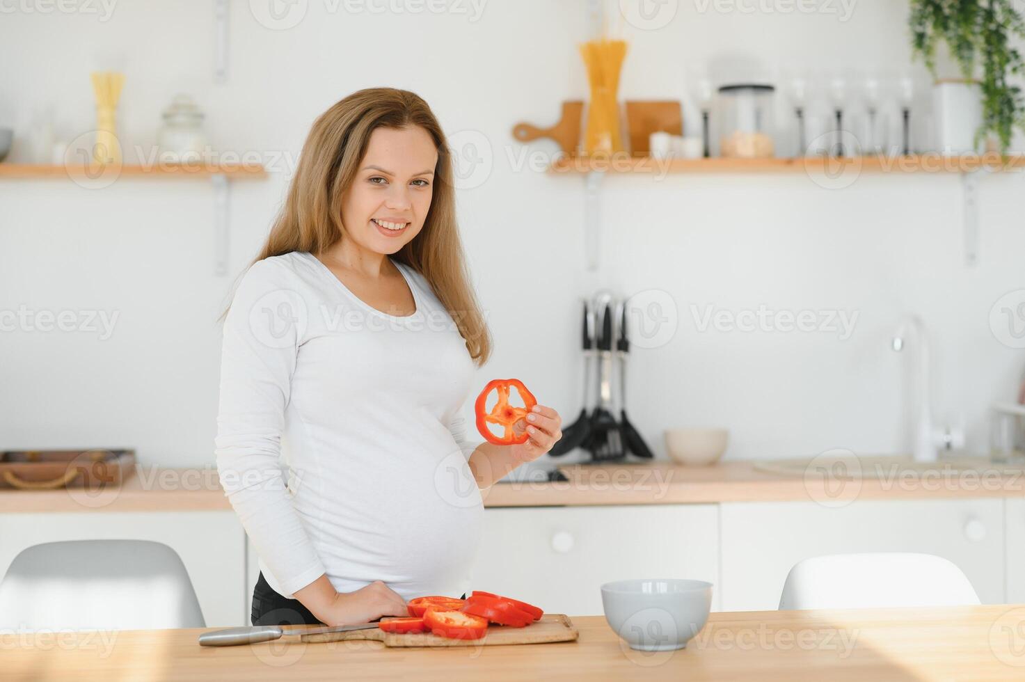 el embarazo, Cocinando alimento, sano estilo de vida, personas y expectativa concepto - contento embarazada mujer con cuchillo el cortar Pepino y preparando vegetal ensalada a hogar foto