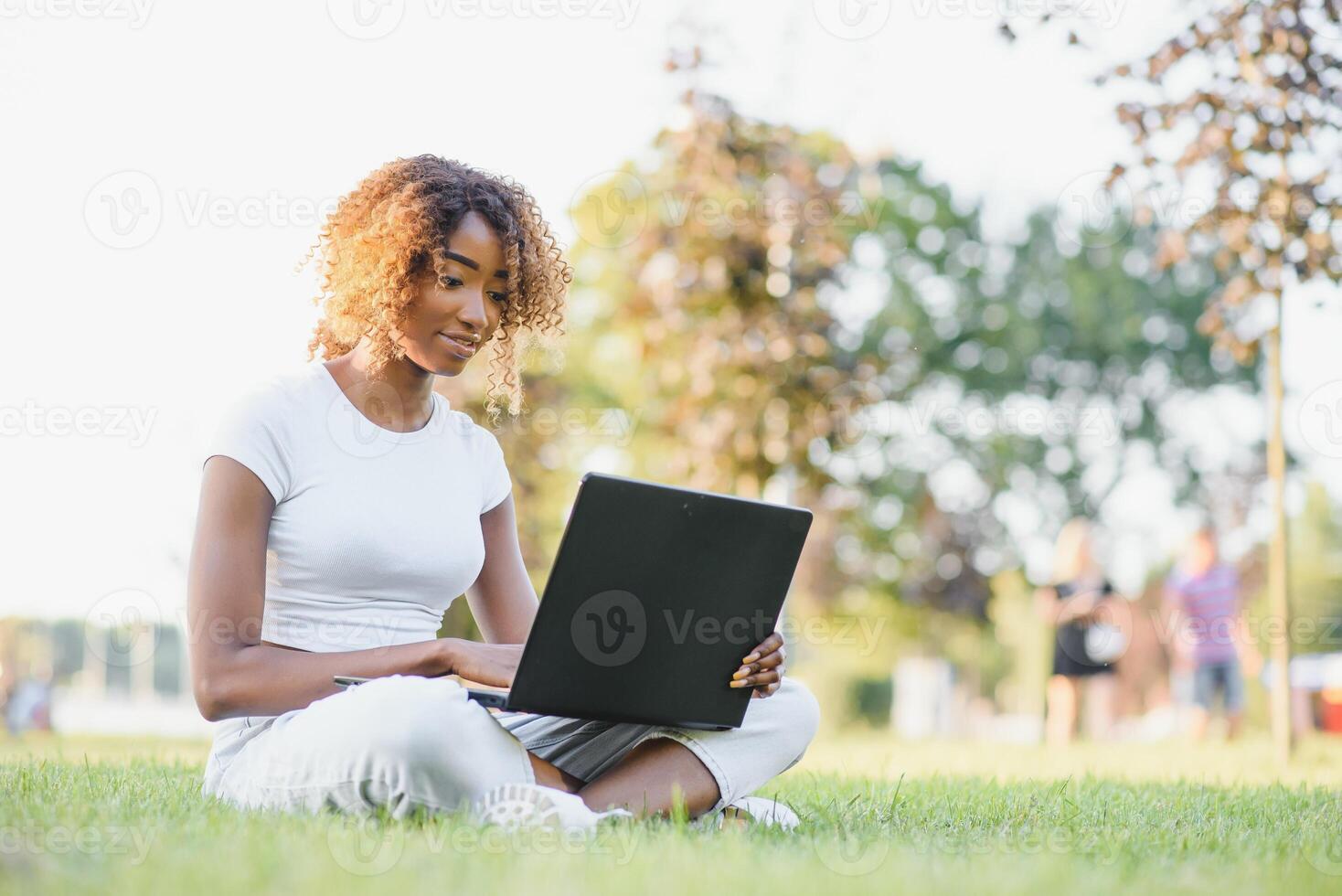 Thoughtful cute mixed female international student with curly hair is sitting on fresh grass with modern laptop in public park, leaning on apple tree and wistfully looking aside during her break photo