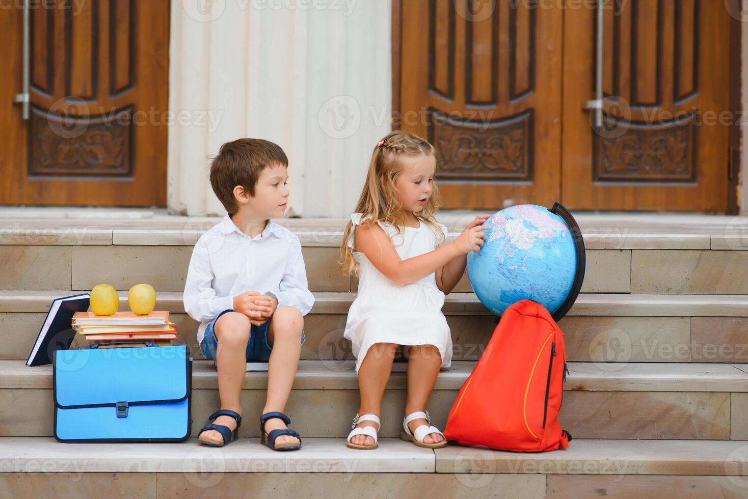 primary education, friendship, childhood and people concept - group of happy elementary school students with backpacks. Two schoolchildren sitting on books near school and eating an apple. photo