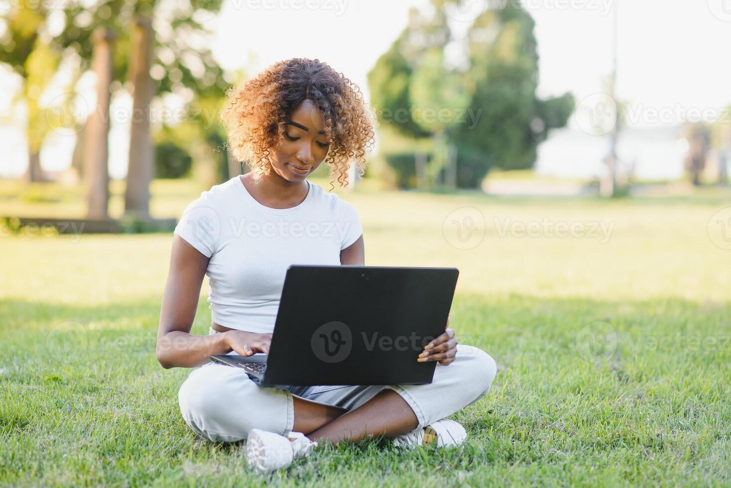 Young attractive dark-skinned college student wearing tank top and jeans sitting on the lawn at campus on sunny day, working on her thesis using laptop computer, looking busy and concentrated photo