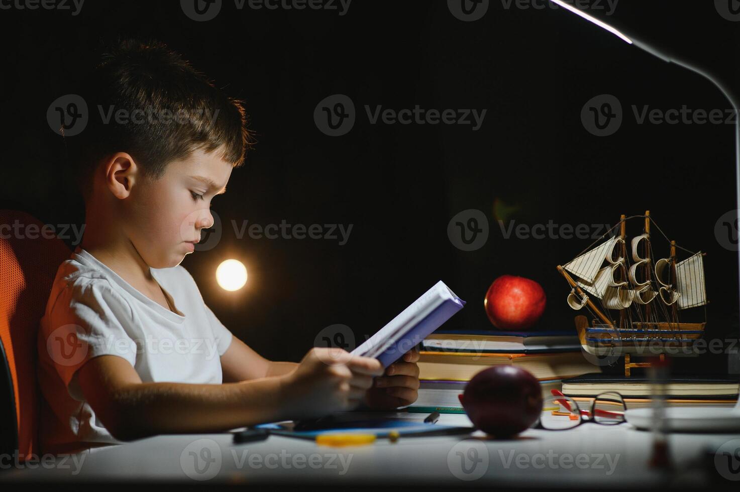 concentrated schoolboy reading book at table with books, plant, lamp, colour pencils, apple, and textbook photo