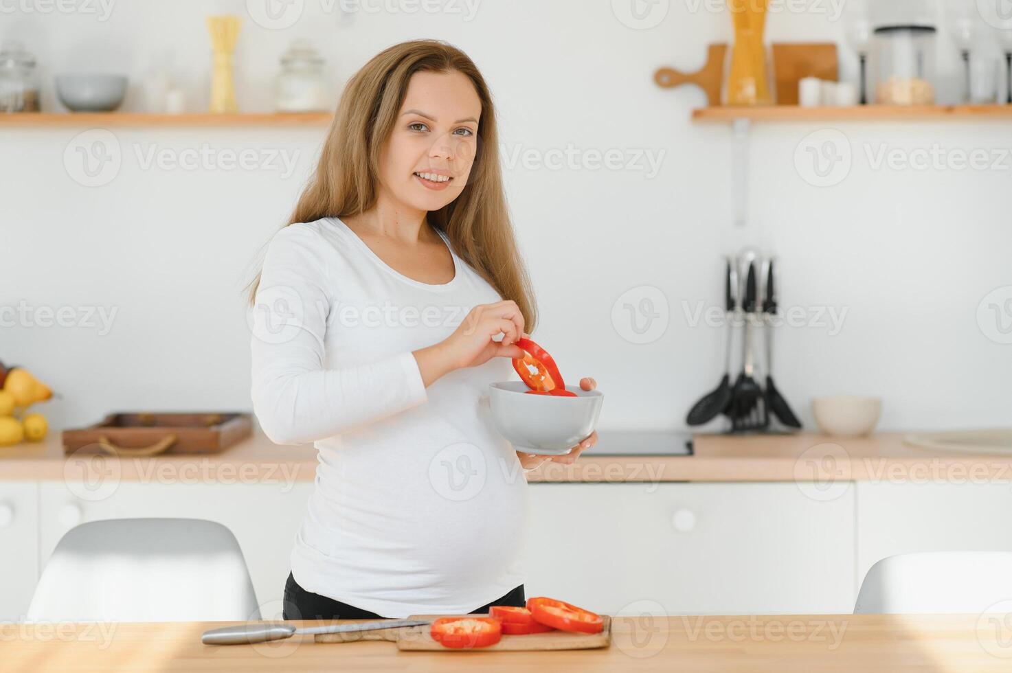 el embarazo, Cocinando alimento, sano estilo de vida, personas y expectativa concepto - contento embarazada mujer con cuchillo el cortar Pepino y preparando vegetal ensalada a hogar foto