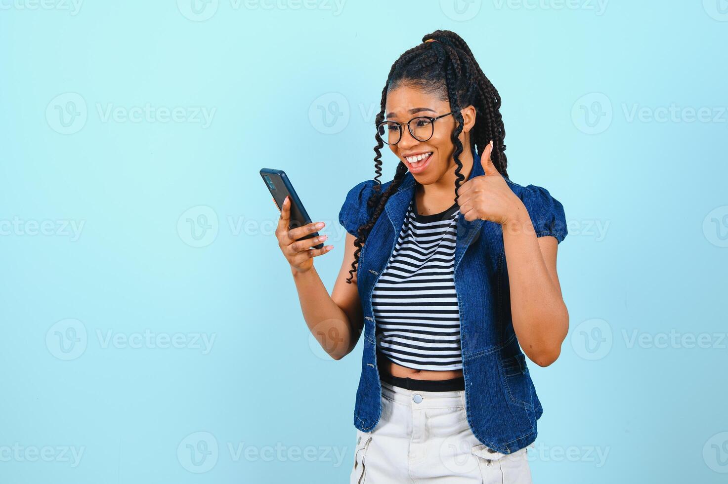 contento alegre afro americano mujer mira a pantalla de inteligente teléfono disfruta en línea chateando tipos texto mensaje surfea social redes vestido por casualidad poses en contra azul antecedentes. tecnología concepto. foto