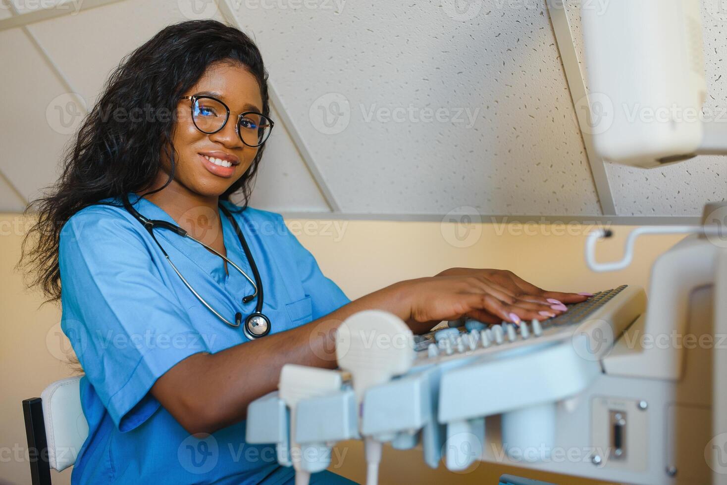 Young female African-american doctor working on modern ultrasound equipment. Operator of ultrasound scanning machine sitting and looking at the monitor, waiting for patient. photo