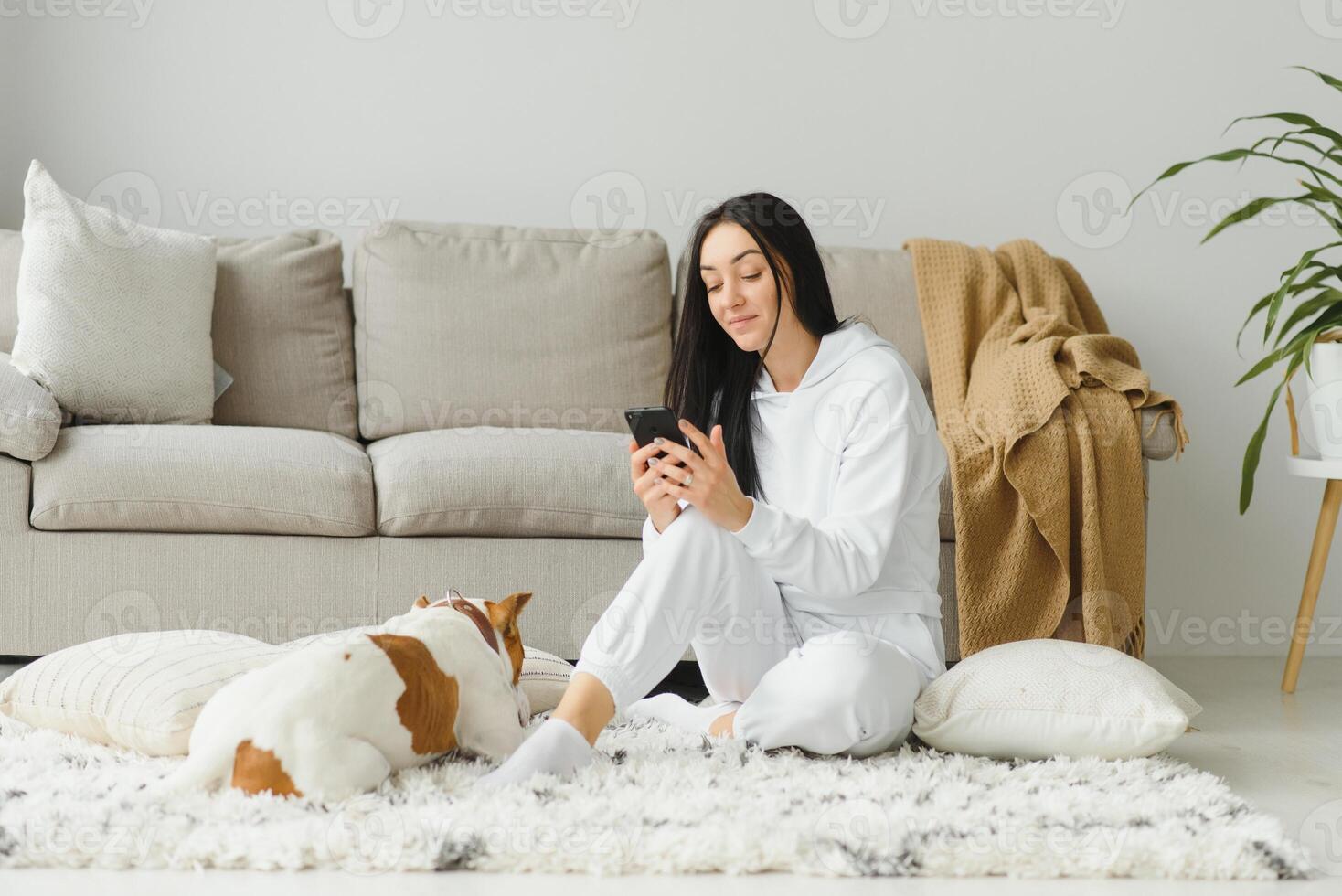 Portrait of beautiful young woman with her dog using mobile phone at home photo
