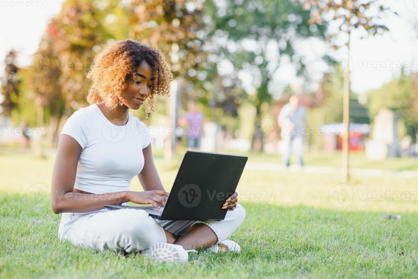 Young attractive dark-skinned college student wearing tank top and jeans sitting on the lawn at campus on sunny day, working on her thesis using laptop computer, looking busy and concentrated photo