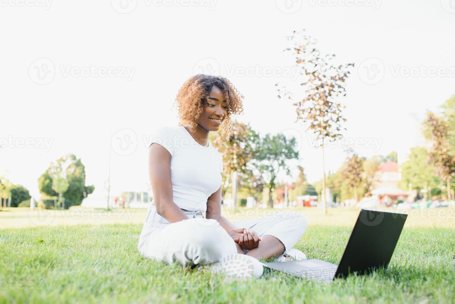 Young attractive dark-skinned college student wearing tank top and jeans sitting on the lawn at campus on sunny day, working on her thesis using laptop computer, looking busy and concentrated photo