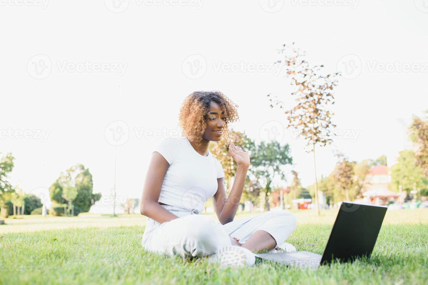 pensativo linda mezclado hembra internacional estudiante con Rizado pelo es sentado en Fresco césped con moderno ordenador portátil en público parque, propensión en manzana árbol y con nostalgia mirando aparte durante su descanso foto